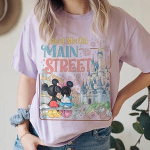 Op de afbeelding: Lichtpaars T-shirt met een kleurrijke afbeelding van Mickey en Minnie Mouse, een kasteel en de tekst "Meet Me On Main Street". Het ontwerp bevat vuurwerk en andere decoratieve elementen.
