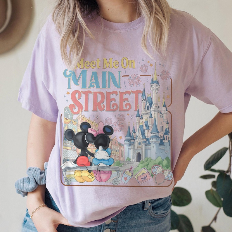 Op de afbeelding: Lichtpaars T-shirt met een kleurrijke afbeelding van Mickey en Minnie Mouse, een kasteel en de tekst "Meet Me On Main Street". Het ontwerp bevat vuurwerk en andere decoratieve elementen.
