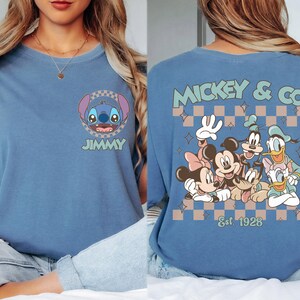 K&ouml;nnte beinhalten: Blaues T-Shirt mit Stitch und dem Namen "JIMMY" auf der Vorderseite. Auf der R&uuml;ckseite steht "MICKEY & CO" mit Disney-Figuren und "Est 1928" in einem Schachbrettmuster. Bequemer, l&auml;ssiger Stil.