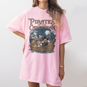 K&ouml;nnte beinhalten: Hellrosa T-Shirt mit einer Grafik von Disney-Figuren in einem Boot und dem Text "Pirates of the Caribbean". Das Design umfasst ein Piratenschiff, einen Totenkopf und einen Nachthimmel mit Vollmond.