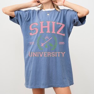 Pu&ograve; includere: T-shirt blu con testo rosa "SHIZ UNIVERSITY". Il design include uno stemma con "EST. 1995". La maglietta ha girocollo e maniche corte. La vestibilit&agrave; &egrave; rilassata.