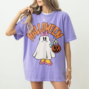 May include: A purple t-shirt with the word "HALLOWEEN" in orange block letters. The shirt features a cartoon ghost character wearing a pink bow, holding a jack-o'-lantern, and wearing pink shoes. The shirt is oversized.
