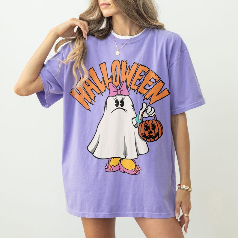 May include: A purple t-shirt with the word "HALLOWEEN" in orange block letters. The shirt features a cartoon ghost character wearing a pink bow, holding a jack-o'-lantern, and wearing pink shoes. The shirt is oversized.