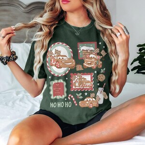 May include: Dark green t-shirt featuring a festive design with gingerbread car illustrations, checkered flags, and the text "HO HO HO." The design includes a candy cane, cookie, and a milk bottle. The shirt has a relaxed fit.