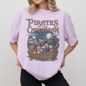 K&ouml;nnte beinhalten: Helllila T-Shirt mit einem Aufdruck von Disney-Figuren in einem Boot und dem Text "Pirates of the Caribbean". Das Design umfasst Micky Maus, Minnie Maus, Goofy und Pluto. Der Hintergrund zeigt ein Piratenschiff, einen Totenkopf und einen Mond.