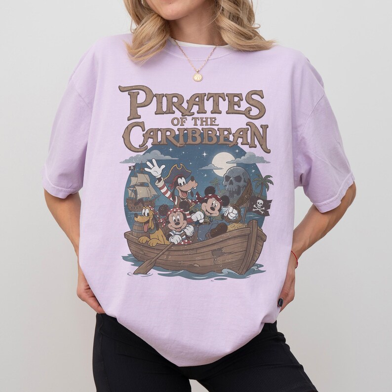 K&ouml;nnte beinhalten: Helllila T-Shirt mit einem Aufdruck von Disney-Figuren in einem Boot und dem Text "Pirates of the Caribbean". Das Design umfasst Micky Maus, Minnie Maus, Goofy und Pluto. Der Hintergrund zeigt ein Piratenschiff, einen Totenkopf und einen Mond.