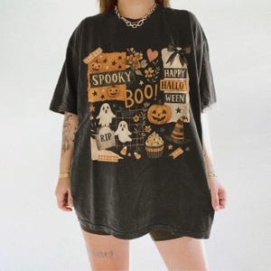 K&ouml;nnte beinhalten: Dunkelgraues T-Shirt mit einem Halloween-Motiv. Das Design umfasst K&uuml;rbisse, Geister, einen Hexenhut und die Aufschriften "Spooky Boo!" und "Happy Halloween." Das T-Shirt hat ein Vintage-Aussehen mit leicht verblichenen Farben.