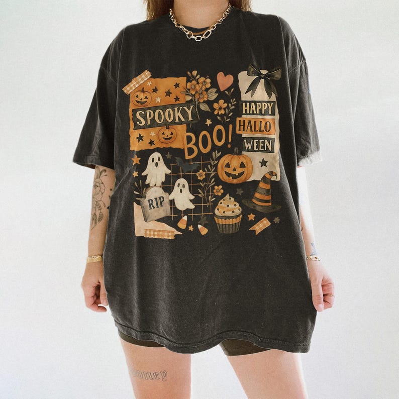 K&ouml;nnte beinhalten: Dunkelgraues T-Shirt mit einem Halloween-Motiv. Das Design umfasst K&uuml;rbisse, Geister, einen Hexenhut und die Aufschriften "Spooky Boo!" und "Happy Halloween." Das T-Shirt hat ein Vintage-Aussehen mit leicht verblichenen Farben.