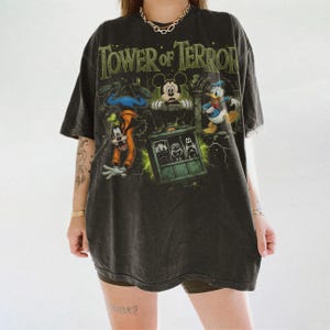 May include: A charcoal gray oversized t-shirt featuring a graphic print of the Tower of Terror. The design includes illustrations of Goofy, Mickey Mouse, and Donald Duck surrounding a central elevator car with green lightning accents and text.