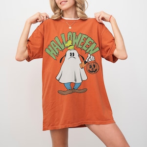 May include: Orange Halloween t-shirt featuring a cartoon character dressed as a ghost, holding a pumpkin-shaped lantern. The word "HALLOWEEN" is printed in green above the character. The shirt has short sleeves and a relaxed fit.