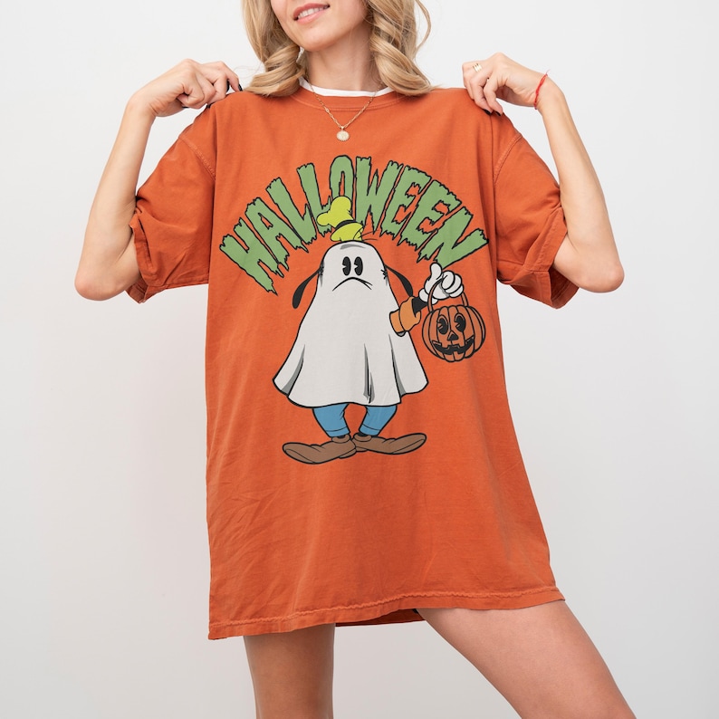 May include: Orange Halloween t-shirt featuring a cartoon character dressed as a ghost, holding a pumpkin-shaped lantern. The word "HALLOWEEN" is printed in green above the character. The shirt has short sleeves and a relaxed fit.