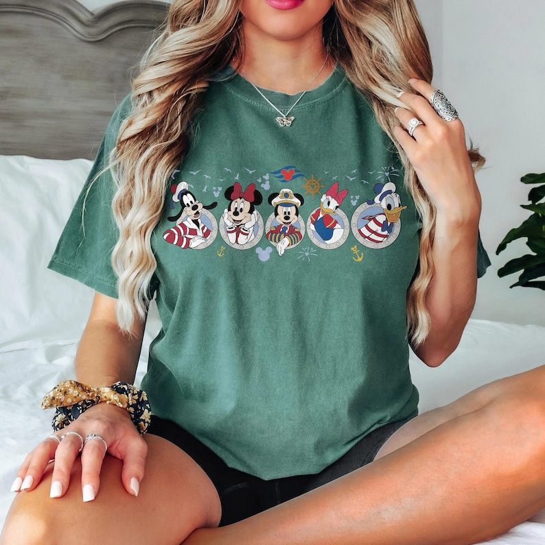 May include: Green t-shirt featuring a row of cartoon characters in circular frames. The characters include Goofy, Minnie Mouse, Mickey Mouse, Daisy Duck, and Donald Duck. The shirt has a crew neck and short sleeves.