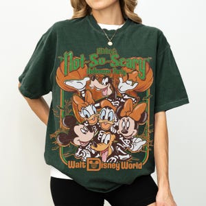 May include: A dark green short-sleeve t-shirt featuring a graphic print of Disney characters in skeleton costumes. The text reads Mickey's Not-So-Scary Halloween Party and Walt Disney World. The design includes pumpkins and bats in orange and green.