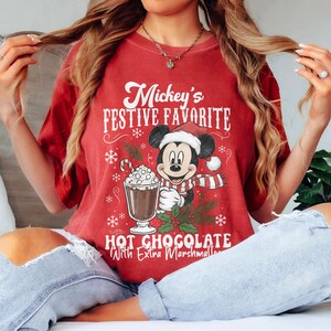 May include: Red t-shirt featuring Mickey Mouse in a Santa hat and scarf, holding a hot chocolate with marshmallows. The text reads "Mickey's Festive Favorite Hot Chocolate With Extra Marshmallows."