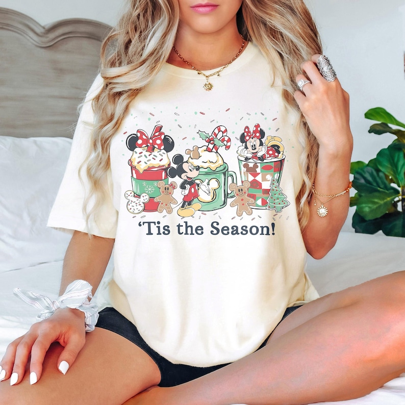 Puede incluir: Camiseta de color crema con un gr&aacute;fico festivo con los personajes de Mickey y Minnie Mouse. El dise&ntilde;o incluye tazas tem&aacute;ticas navide&ntilde;as, hombres de jengibre y el texto "'Tis the Season!" Una camiseta divertida y de temporada para los fans de Disney.