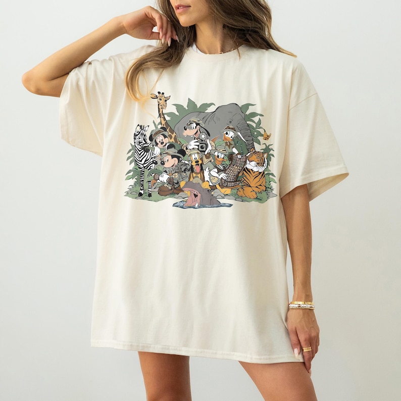 K&ouml;nnte beinhalten: Cremefarbenes, &uuml;bergro&szlig;es T-Shirt mit einem farbenfrohen Grafikdesign von Zeichentierfiguren, darunter ein Zebra, eine Giraffe, ein Elefant, ein Tiger und Mickey Mouse. Das Design ist vor einem Hintergrund aus gr&uuml;nem Laub angeordnet.