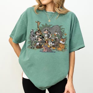 K&ouml;nnte beinhalten: Gr&uuml;nes T-Shirt mit einem Cartoon-Motiv, das verschiedene Tiere zeigt, darunter ein Zebra, eine Giraffe, ein Elefant, ein Tiger und ein Nilpferd, in einer Dschungelszene. Das T-Shirt hat einen Rundhalsausschnitt und kurze &Auml;rmel.