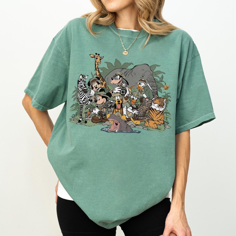 K&ouml;nnte beinhalten: Gr&uuml;nes T-Shirt mit einem Cartoon-Motiv, das verschiedene Tiere zeigt, darunter ein Zebra, eine Giraffe, ein Elefant, ein Tiger und ein Nilpferd, in einer Dschungelszene. Das T-Shirt hat einen Rundhalsausschnitt und kurze &Auml;rmel.