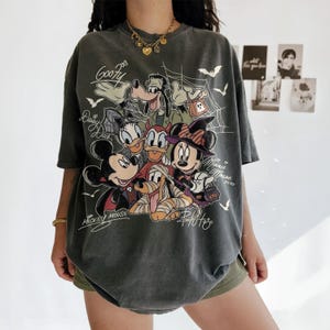 Może przedstawiać: Ciemnoszary t-shirt z kr&oacute;tkim rękawem z grafiką o tematyce Halloween przedstawiającą przebrane postacie Disneya, takie jak Myszka Miki, Myszka Minnie, Goofy, Kaczka Daisy i Pluto, z napisami przy każdej postaci oraz motywami pajęczyn.