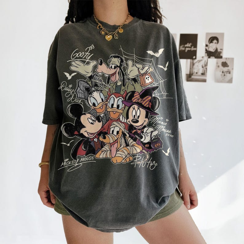 Może przedstawiać: Ciemnoszary t-shirt z kr&oacute;tkim rękawem z grafiką o tematyce Halloween przedstawiającą przebrane postacie Disneya, takie jak Myszka Miki, Myszka Minnie, Goofy, Kaczka Daisy i Pluto, z napisami przy każdej postaci oraz motywami pajęczyn.
