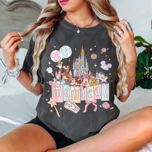 May include: Dark gray t-shirt featuring a colorful graphic with Disney characters, a castle, and the word "magical". The design includes Mickey Mouse, Minnie Mouse, Donald Duck, and Goofy. Pink and blue accents are used throughout the design.