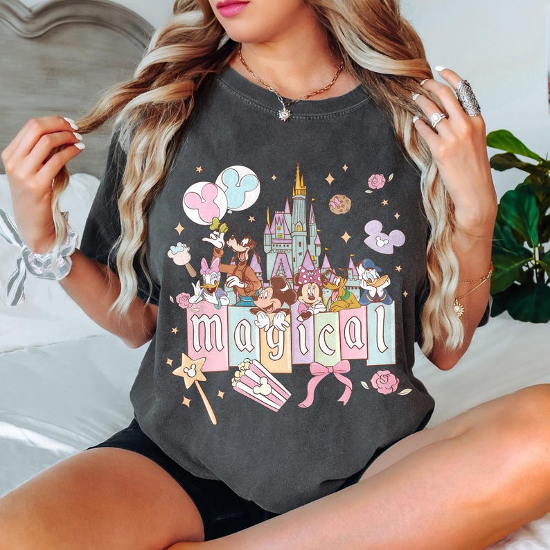 May include: Dark gray t-shirt featuring a colorful graphic with Disney characters, a castle, and the word "magical". The design includes Mickey Mouse, Minnie Mouse, Donald Duck, and Goofy. Pink and blue accents are used throughout the design.