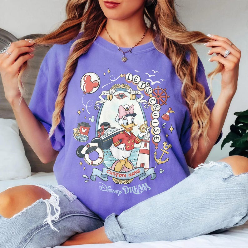 K&ouml;nnte beinhalten: Ein lila T-Shirt mit einer Grafik von Daisy Duck in Kapit&auml;nsuniform, einem Kreuzfahrtschiff und dem Text "Let's Cruise 2024". Das Design enth&auml;lt nautische Elemente wie einen Leuchtturm, einen Anker und ein Steuerrad. Das Shirt hat auch den Text "Disney Dream".