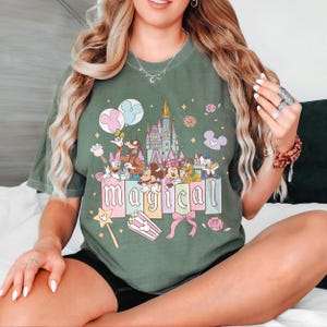 May include: Olive green t-shirt featuring a colorful graphic of Disney characters, a castle, and the word "magical". The design includes Mickey and Minnie Mouse, Donald Duck, and other characters. The shirt has a crew neck and short sleeves.