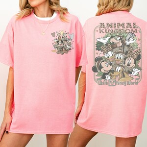 Puede incluir: Camiseta rosa claro con un dise&ntilde;o de Disney. La parte delantera tiene un peque&ntilde;o dise&ntilde;o con Mickey y Minnie Mouse, y la parte trasera muestra "ANIMAL KINGDOM" y "Walt Disney World". Los personajes visten trajes de safari.