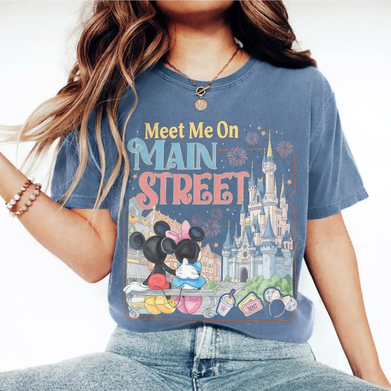 Op de afbeelding: Blauw T-shirt met de tekst "Meet Me On Main Street" en een afbeelding van Mickey en Minnie Mouse voor een kasteel. Het ontwerp bevat vuurwerk, een drankje en muisoren-haarbanden. Het shirt is een comfortabele, casual stijl.