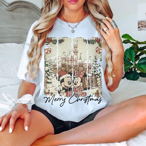 May include: White t-shirt featuring a distressed graphic of Mickey and Minnie Mouse in Christmas attire, with a castle backdrop and the words "Merry Christmas". The design includes candy canes and Christmas trees.