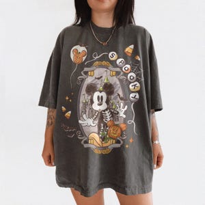 May include: Dark gray oversized t-shirt featuring a Halloween-themed graphic. The design includes a skeleton Mickey Mouse, a pumpkin, and the word "Spooky". Additional elements include a castle, bats, and candy corn.