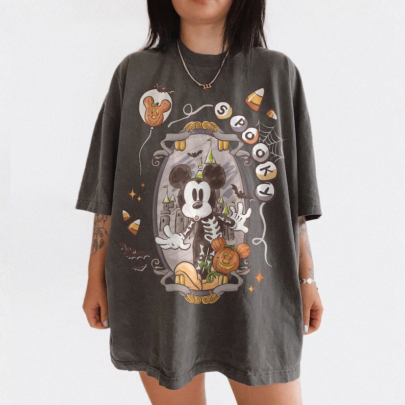 May include: Dark gray oversized t-shirt featuring a Halloween-themed graphic. The design includes a skeleton Mickey Mouse, a pumpkin, and the word "Spooky". Additional elements include a castle, bats, and candy corn.