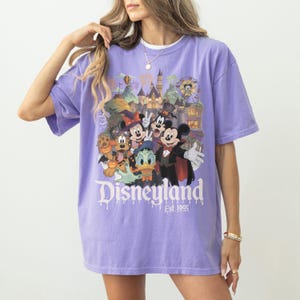 May include: A light purple short-sleeve t-shirt featuring a graphic print of Disney characters in Halloween costumes, including Mickey Mouse, Minnie Mouse, Donald Duck, and Pluto. The text Disneyland Est. 1955 is printed below the characters.