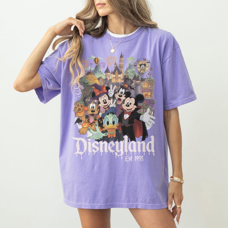 May include: A light purple short-sleeve t-shirt featuring a graphic print of Disney characters in Halloween costumes, including Mickey Mouse, Minnie Mouse, Donald Duck, and Pluto. The text Disneyland Est. 1955 is printed below the characters.