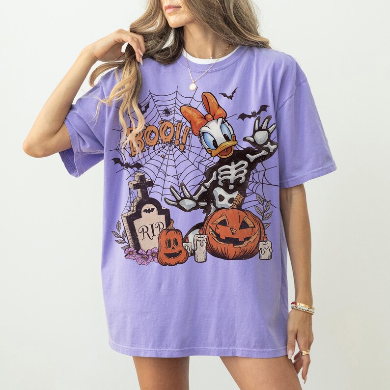 May include: A light purple short-sleeve t-shirt featuring a graphic print of a cartoon duck in a skeleton costume, a spider web, a tombstone with the text RIP, a pumpkin, and the word BOO. The design includes bats and candles.