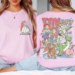 K&ouml;nnte beinhalten: Hellrosa T-Shirt mit einer Prinzessinnen-Grafik und dem Wort "Auntie" auf der Vorderseite. Auf der R&uuml;ckseite des Shirts stehen die Worte "Toy Story" und Charaktere wie Woody, Buzz und Rex.