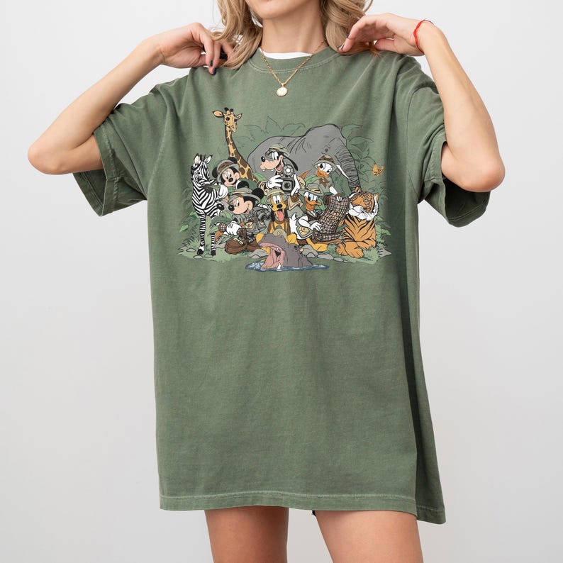 K&ouml;nnte beinhalten: Olivgr&uuml;nes T-Shirt mit einem Safari-Motiv. Das Design zeigt Zeichentricktiere wie Zebra, Giraffe, Elefant und Tiger sowie Micky Maus und Freunde, alle in Safari-Outfits.