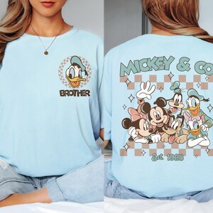 K&ouml;nnte beinhalten: Hellblaues T-Shirt mit Donald Duck und dem Wort "Brother". Auf der R&uuml;ckseite Mickey & Co. mit Disney-Figuren und dem Text "Est. 1928". Das Design hat ein Schachbrettmuster.