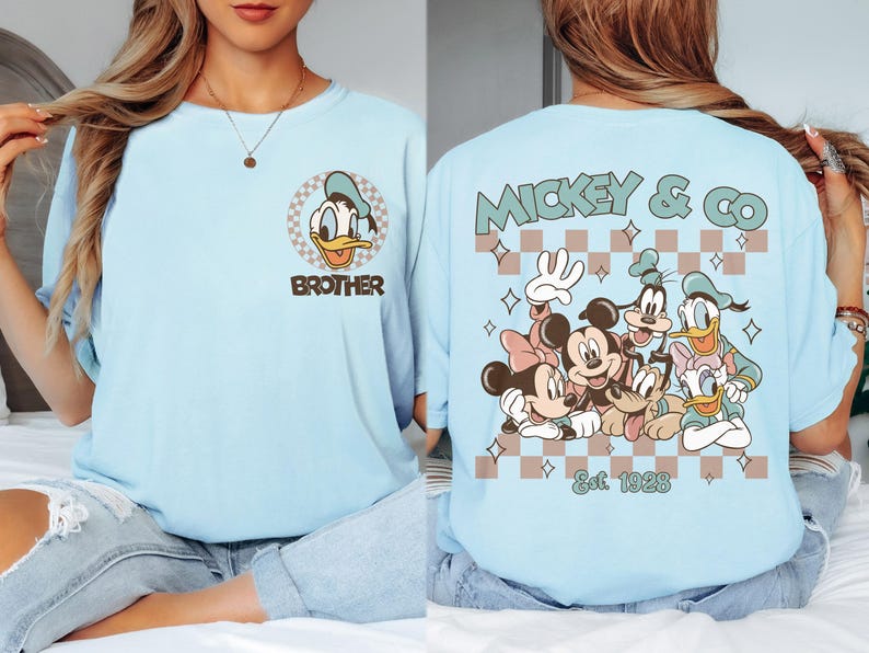 K&ouml;nnte beinhalten: Hellblaues T-Shirt mit Donald Duck und dem Wort "Brother". Auf der R&uuml;ckseite Mickey & Co. mit Disney-Figuren und dem Text "Est. 1928". Das Design hat ein Schachbrettmuster.