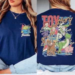 K&ouml;nnte beinhalten: Marineblaues T-Shirt mit einem Cartoon-Alien und dem Namen "Justin" auf der Vorderseite. Auf der R&uuml;ckseite befindet sich eine "Toy Story"-Grafik mit Figuren wie Woody, Buzz und Rex. Das Design enth&auml;lt den Text "Jessie Since 1995".