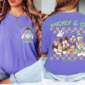 K&ouml;nnte beinhalten: Lilafarbenes T-Shirt mit einem Cartoon-Eeyore-Motiv und dem Namen "LAURA" auf der Vorderseite. Auf der R&uuml;ckseite ist eine Disney-Figurengruppe mit "MICKEY & CO" und "Est 1928" abgebildet.