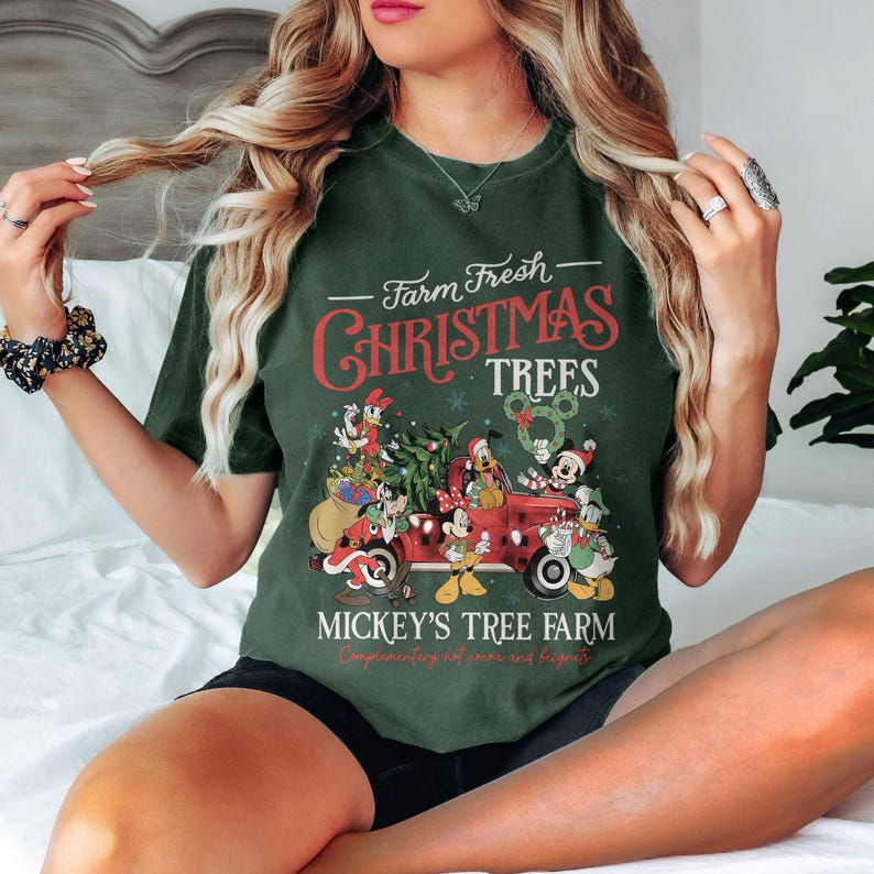 May include: Dark green t-shirt with a Christmas-themed design. The shirt features the text "Farm Fresh Christmas Trees" above a red truck filled with Disney characters and a Christmas tree. Below, it reads "Mickey's Tree Farm".