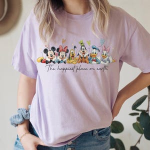 May include: A light purple t-shirt featuring a graphic of Disney characters Mickey Mouse, Minnie Mouse, Pluto, Goofy, Donald Duck, and Daisy Duck. The text "The happiest place on earth" is written below the characters.