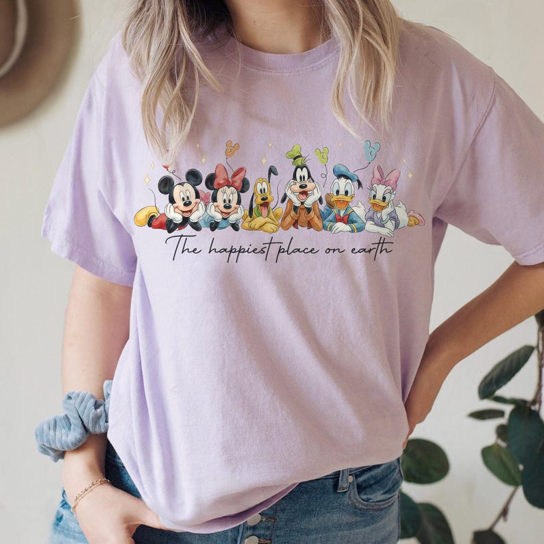 May include: A light purple t-shirt featuring a graphic of Disney characters Mickey Mouse, Minnie Mouse, Pluto, Goofy, Donald Duck, and Daisy Duck. The text "The happiest place on earth" is written below the characters.
