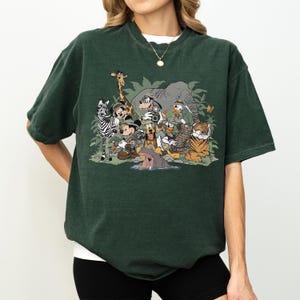 K&ouml;nnte beinhalten: Dunkelgr&uuml;nes T-Shirt mit einem farbenfrohen Grafikdesign von Zeichentrickfiguren in einer Dschungelkulisse. Das Design umfasst ein Zebra, eine Giraffe, einen Elefanten, einen Tiger und andere Tiere sowie mehrere Zeichentrickfiguren. Das T-Shirt hat kurze &Auml;rmel und einen Rundhalsausschnitt.