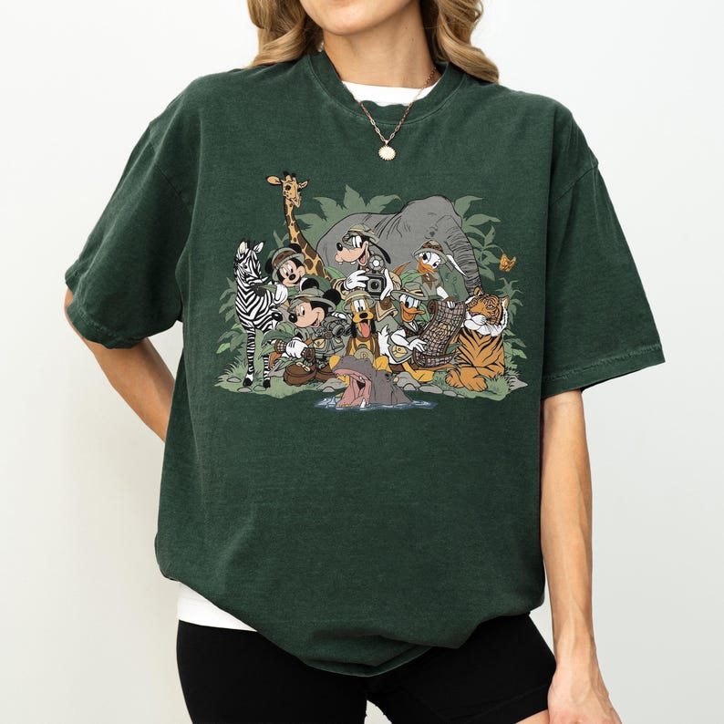 K&ouml;nnte beinhalten: Dunkelgr&uuml;nes T-Shirt mit einem farbenfrohen Grafikdesign von Zeichentrickfiguren in einer Dschungelkulisse. Das Design umfasst ein Zebra, eine Giraffe, einen Elefanten, einen Tiger und andere Tiere sowie mehrere Zeichentrickfiguren. Das T-Shirt hat kurze &Auml;rmel und einen Rundhalsausschnitt.
