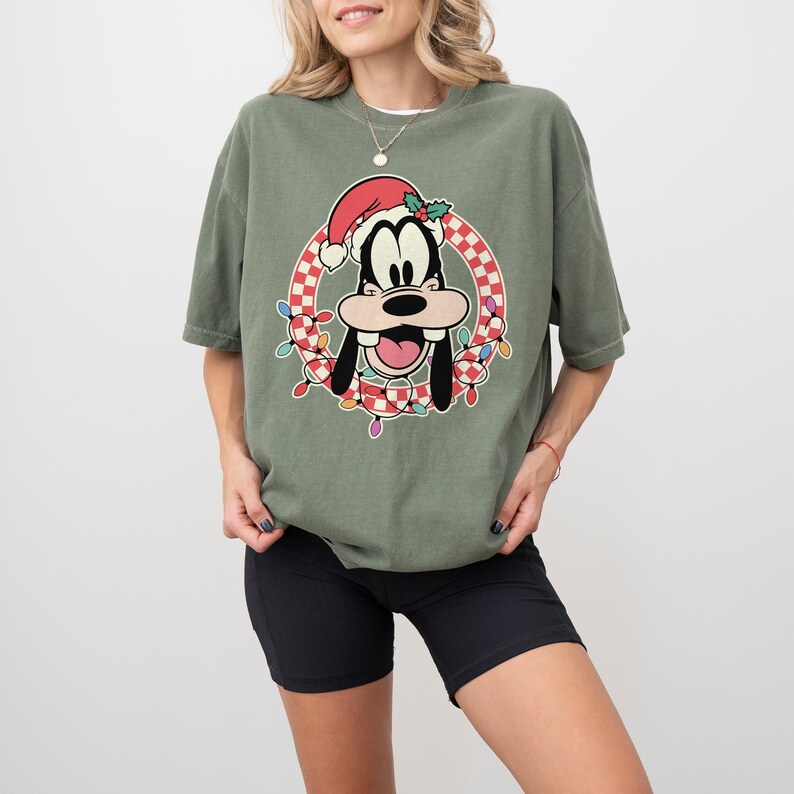 May include: Olive green t-shirt featuring a cartoon image of Goofy wearing a Santa hat and surrounded by a wreath of Christmas lights. The graphic is set against a red and white checkered background. The shirt has short sleeves.