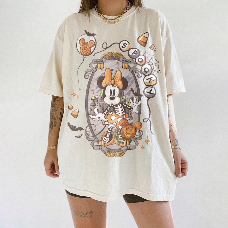 May include: Cream-colored t-shirt featuring a Halloween-themed Minnie Mouse design. The graphic includes a skeleton Minnie, a pumpkin, bats, and the words "Spotlight" and "Minnie Mouse". The shirt has short sleeves and a relaxed fit.