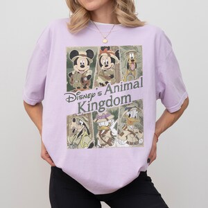 May include: A light purple t-shirt featuring a graphic of Disney's Animal Kingdom characters. The design includes Mickey Mouse, Minnie Mouse, Pluto, Goofy, and Donald Duck in safari outfits. The text "Disney's Animal Kingdom" is also displayed.